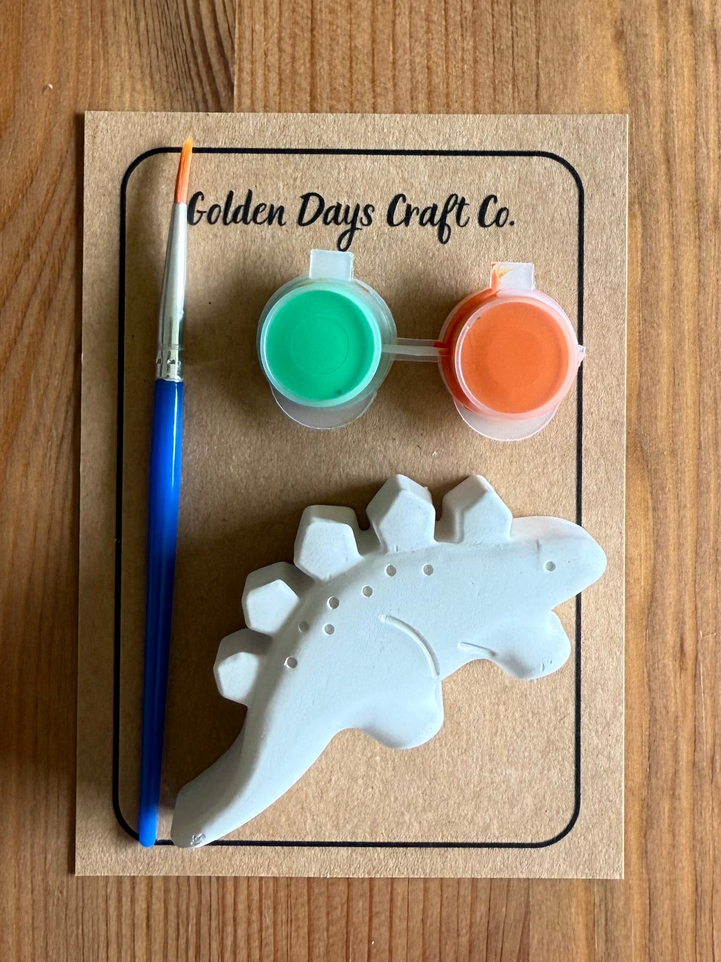 A dinosaur-shaped ceramic painting kit by Golden Days Craft Co. on a wooden background, featuring a white unpainted dinosaur, a blue-handled paintbrush, two small paint pots (green and orange), and a kraft paper backing card.