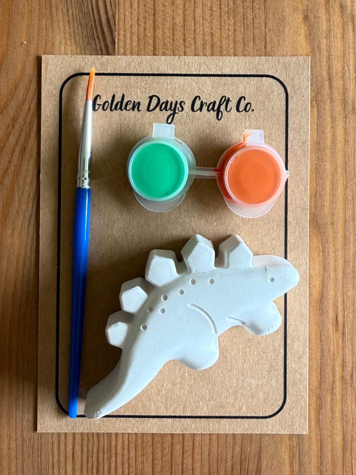 A dinosaur-shaped ceramic painting kit by Golden Days Craft Co. on a wooden background, featuring a white unpainted dinosaur, a blue-handled paintbrush, two small paint pots (green and orange), and a kraft paper backing card.
